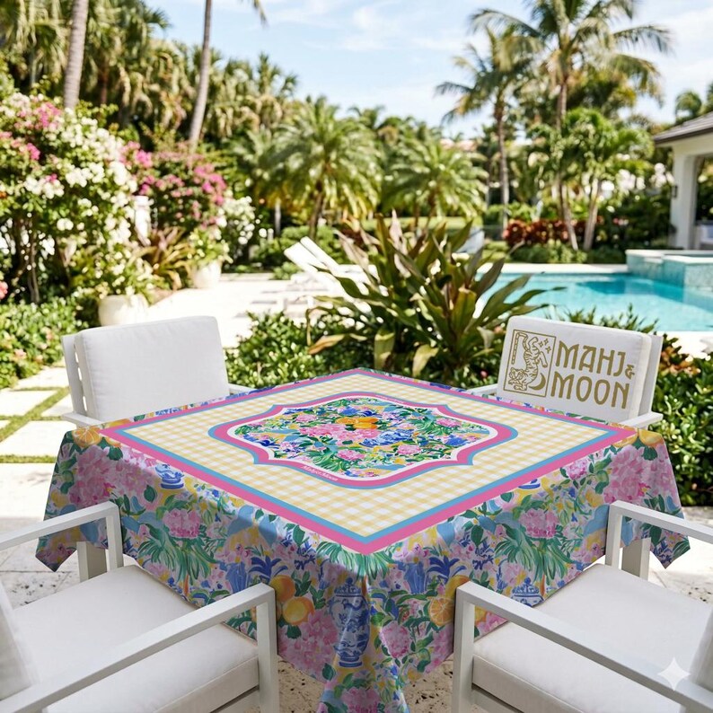 May include: A square tablecloth featuring a central floral pattern with pink, blue, and yellow flowers, surrounded by a yellow and white gingham border and a pink frame. The sides display a repeating print of blue and white vases and flowers.