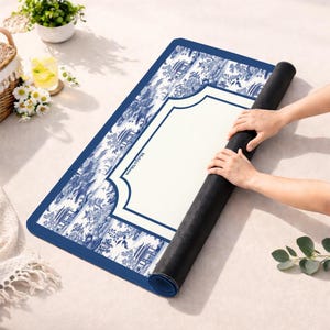 May include: A rectangular mat featuring a blue and white toile pattern with illustrations of pagodas and trees. The center has a solid cream-colored rectangular area with the text Mahjong Moon printed in black. The underside is a solid black material.
