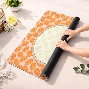 May include: A rolled-up yoga mat with a pumpkin pattern. The mat features a light peach background with orange pumpkins. A circular design in the center has a light green border and the words "MAHI + MOON". The mat is black on the underside.