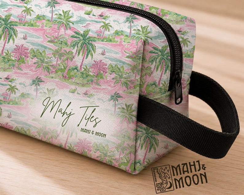 Op de afbeelding: Een rechthoekige toilettas met een tropische eilandprint in roze, groen en wit. De tas heeft een zwarte rits, een zwart handvat en de tekst "Mahj Tiles" in groen script. Het merklogo "Mahj & Moon" is ook zichtbaar.
