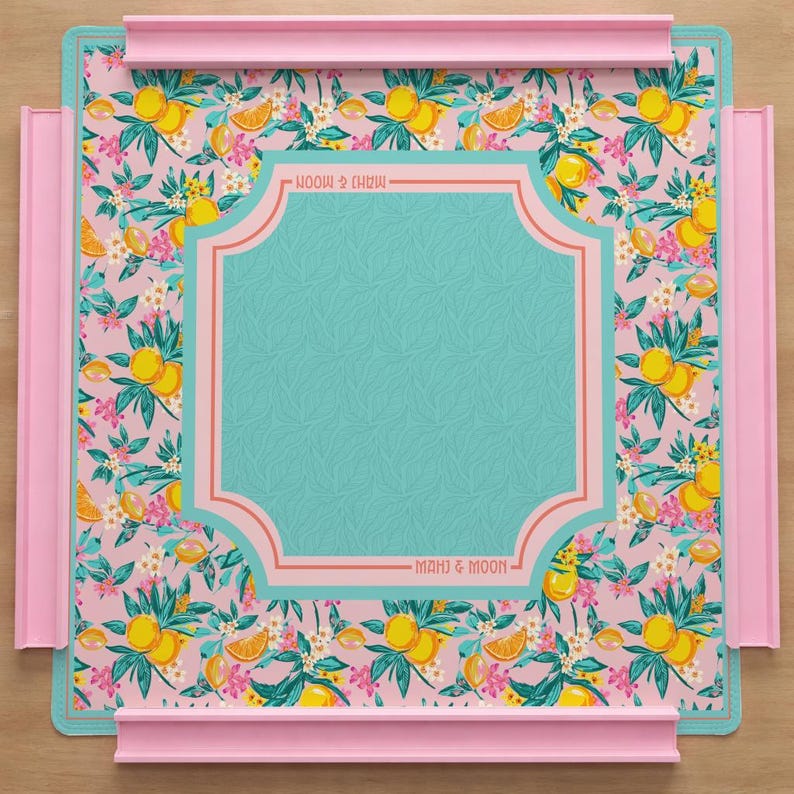 May include: A square mahjong mat featuring a pink background with a repeating pattern of yellow lemons, green leaves, and small white and pink flowers. A central teal panel with a subtle leaf texture is framed by pink lines with the text MAHJ & MOON.