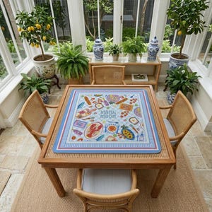 May include: A square mahjong mat featuring a light blue and white checkered pattern with a border of blue and red stripes. The mat displays colorful illustrations of food items including a lobster, bread, and wine, with the text "MAHJ MOON" in the center.