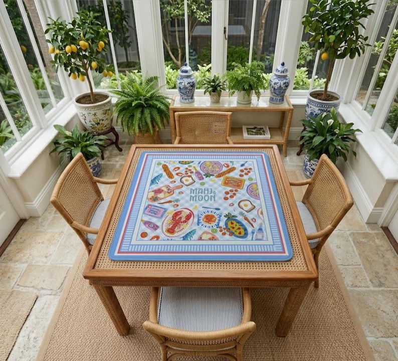 May include: A square mahjong mat featuring a light blue and white checkered pattern with a border of blue and red stripes. The mat displays colorful illustrations of food items including a lobster, bread, and wine, with the text "MAHJ MOON" in the center.