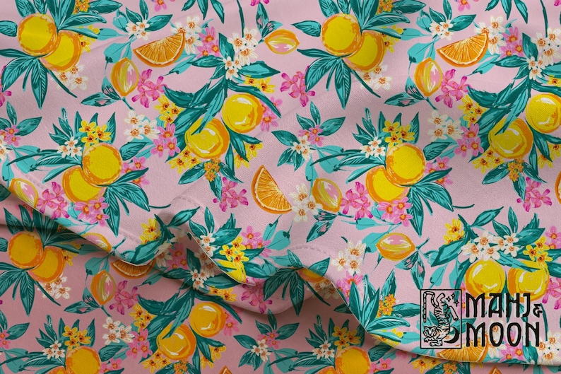 May include: A fabric with a repeating pattern of yellow lemons, orange slices, and white and pink flowers on a light pink background. The leaves are teal and the design is from MAHI & MOON.