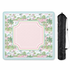 Op de afbeelding: Een vierkante, lichtblauwe mat met een tropisch ontwerp met roze flamingo's, palmbomen en een roze en goudkleurig omlijst midden. Een zwarte draagtas staat naast de mat. De mat heeft de tekst "Mahly Moon" in de hoek.