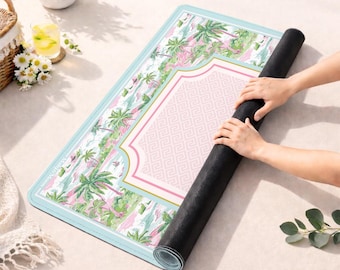 May include: A rectangular exercise mat featuring a light blue border and a central pink geometric pattern. The surrounding area displays a tropical print with green palm trees and pink accents. The mat is partially rolled, revealing a black underside.