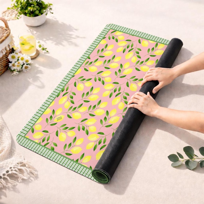 May include: A rectangular mat featuring a pink background with a repeating pattern of yellow lemons and green leaves. The mat has a dark green and white striped border. The underside of the mat is solid black, and it is partially rolled up.