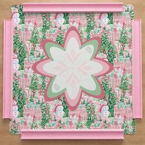 Op de afbeelding: Een vierkant puzzelbord met een roze frame met een herhalend patroon van roze huizen, groene dennenbomen en sneeuwpoppen. In het midden toont een groot, meerlaags, stervormig stuk een wit, roze en groen bloemachtig ontwerp.