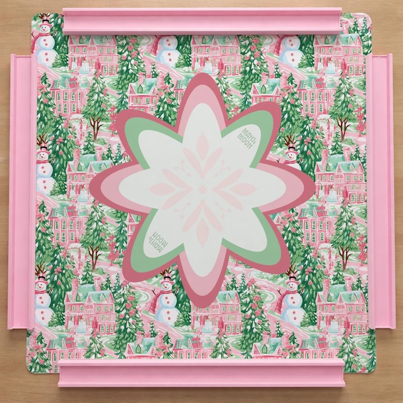 Op de afbeelding: Een vierkant puzzelbord met een roze frame met een herhalend patroon van roze huizen, groene dennenbomen en sneeuwpoppen. In het midden toont een groot, meerlaags, stervormig stuk een wit, roze en groen bloemachtig ontwerp.