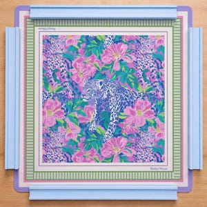 May include: A square Mahjong mat featuring a vibrant pattern of leopards and pink flowers on a green background. The mat is framed by a light blue plastic border with four raised edges. The text Mahjo Moon is printed in the corners of the mat.