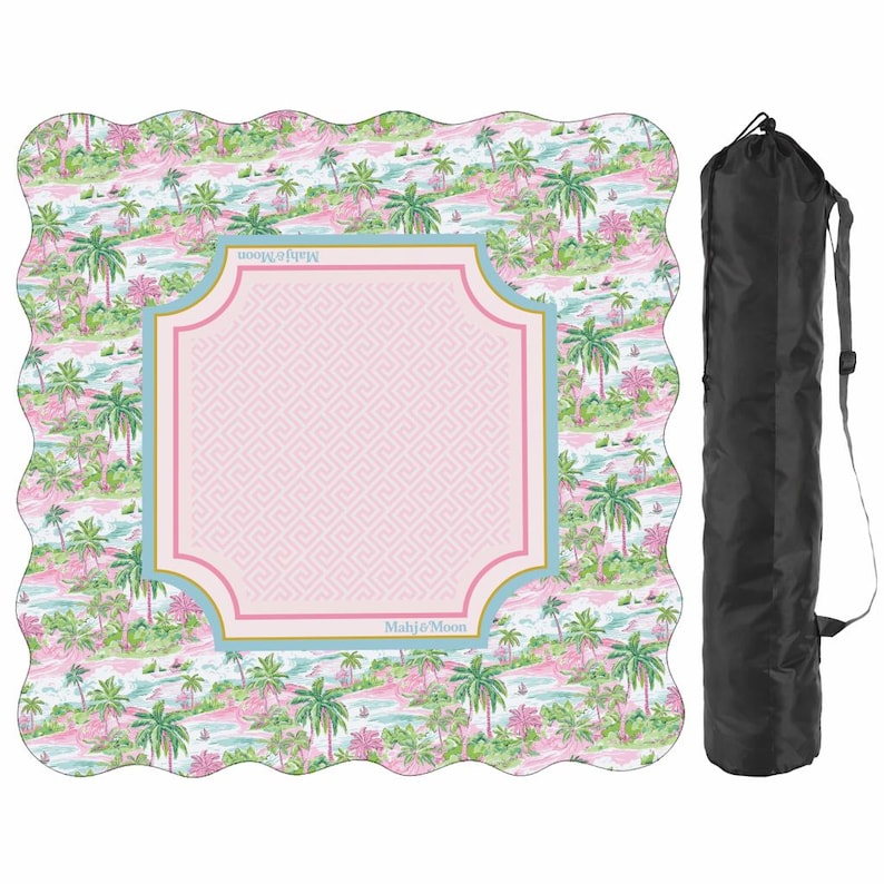 May include: A square mat with scalloped edges featuring a pink and green tropical palm tree pattern and a central light pink square with a Greek key border. The text Mahj&Moon is printed on the border. A black cylindrical carrying bag is included.