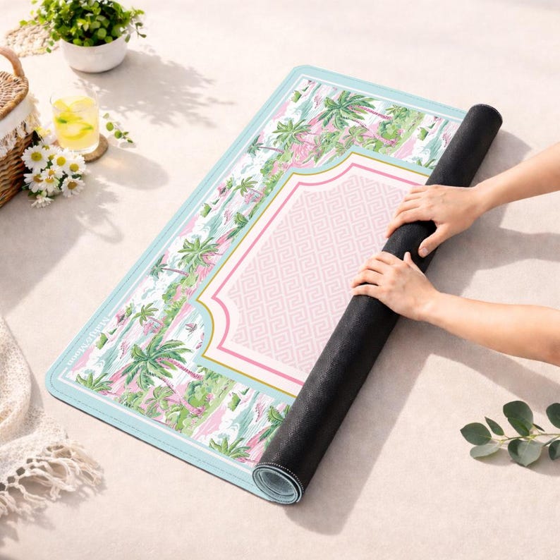 Op de afbeelding: Een opgerolde yogamat met een tropisch ontwerp. De mat heeft een lichtblauwe rand, een roze midden met een Grieks sleutelpatroon en een tropische print met palmbomen in groen en roze. De mat wordt uitgerold.