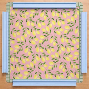 May include: A square mat featuring a repeating pattern of yellow lemons and green leaves on a pink background, framed by a thin green and white striped border. The mat is held in place by four light blue plastic bars on each side.
