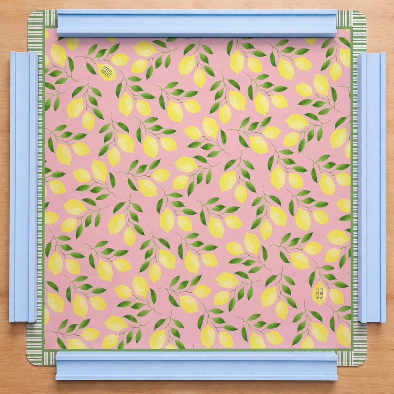 May include: A square mat featuring a repeating pattern of yellow lemons and green leaves on a pink background, framed by a thin green and white striped border. The mat is held in place by four light blue plastic bars on each side.