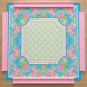 Op de afbeelding: Een vierkant spelbord met een levendig tropisch palmbladpatroon in roze, blauw en groen. Het midden heeft een lichtgroen geschulpt ontwerp omlijst door roze en witte randen. De tekst "MAHJ & MOON" staat op het bord, dat is omringd door vier verhoogde roze plastic wanden.