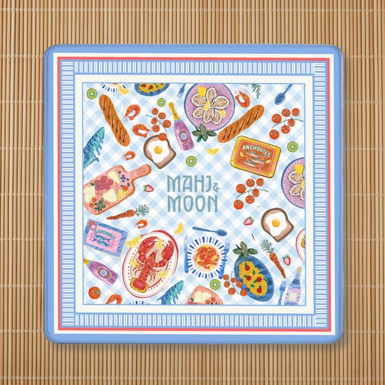 May include: A square textile with a light blue and white gingham pattern featuring illustrations of food items including a lobster, oysters, bread, anchovies, and vegetables. The text MAHJ & MOON is centered in blue. The border is striped blue and white.