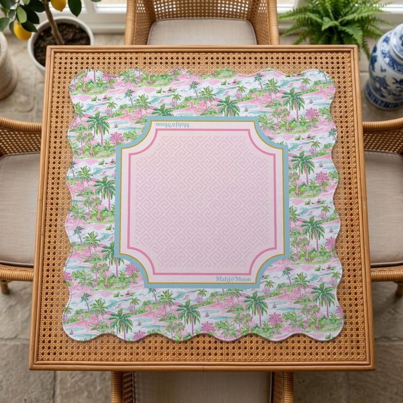 May include: A square scalloped-edge mat featuring a tropical palm tree pattern in shades of pink and green. The centre displays a light pink square with a Greek key border, outlined in blue and pink, with the text Mahjong Moon printed on it.