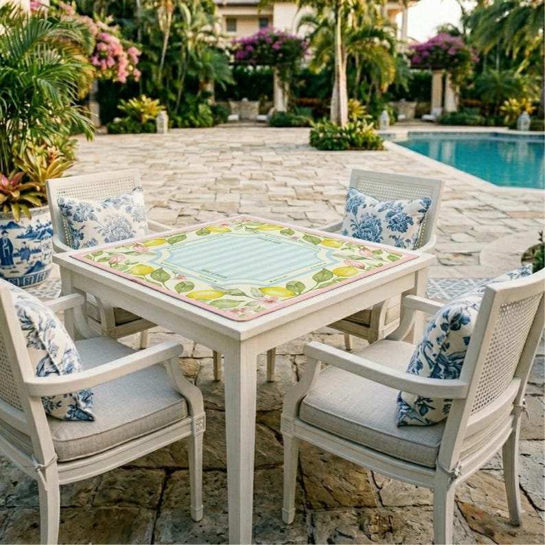 K&ouml;nnte beinhalten: Ein wei&szlig;er quadratischer Gartentisch mit vier passenden St&uuml;hlen. Der Tisch hat eine Tischdecke mit Zitronenmotiv und einem hellblau gestreiften Mittelteil. Die St&uuml;hle haben hellgraue Kissen und blau gebl&uuml;mte Kissen. Die Szene spielt sich im Freien in der N&auml;he eines Pools ab.