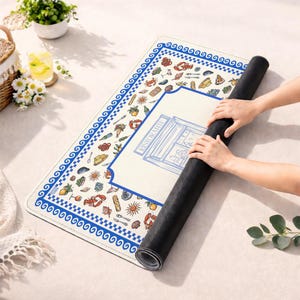 Puede incluir: Una alfombrilla rectangular con superficie color crema y un borde griego azul y blanco. El dise&ntilde;o incluye ilustraciones coloridas de mariscos, frutas y cubiertos, con un gr&aacute;fico central de l&iacute;neas azules etiquetado como RISTORANTE.