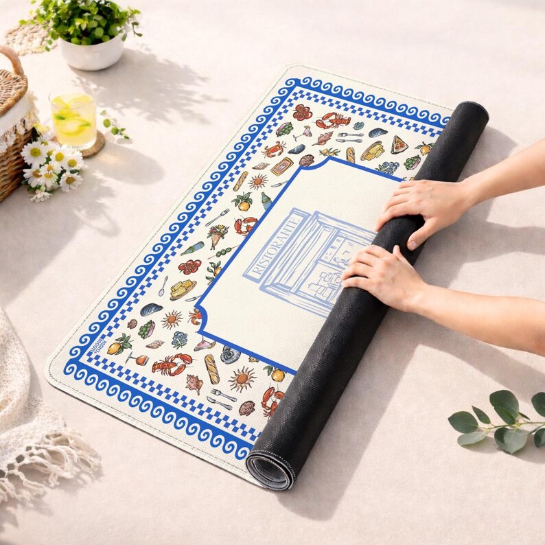 Puede incluir: Una alfombrilla rectangular con superficie color crema y un borde griego azul y blanco. El dise&ntilde;o incluye ilustraciones coloridas de mariscos, frutas y cubiertos, con un gr&aacute;fico central de l&iacute;neas azules etiquetado como RISTORANTE.