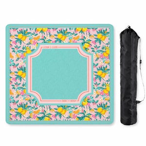 May include: A square picnic blanket featuring a pink background with a repeating pattern of yellow lemons, green leaves, and white flowers, centered by a teal panel with a pink border and the text "MAHI & MOON" and a black carrying bag.
