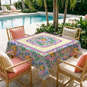 May include: A square tablecloth featuring a vibrant floral and citrus pattern with lemons and pink blossoms, bordered by a yellow and white gingham check. The center displays a decorative medallion, and the fabric drapes over a square table.