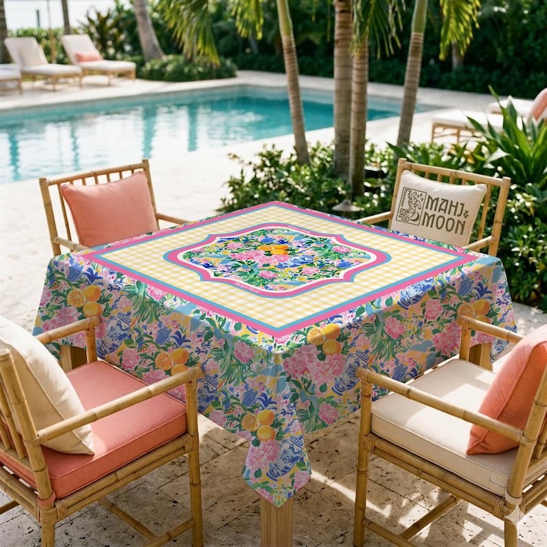 May include: A square tablecloth featuring a vibrant floral and citrus pattern with lemons and pink blossoms, bordered by a yellow and white gingham check. The center displays a decorative medallion, and the fabric drapes over a square table.