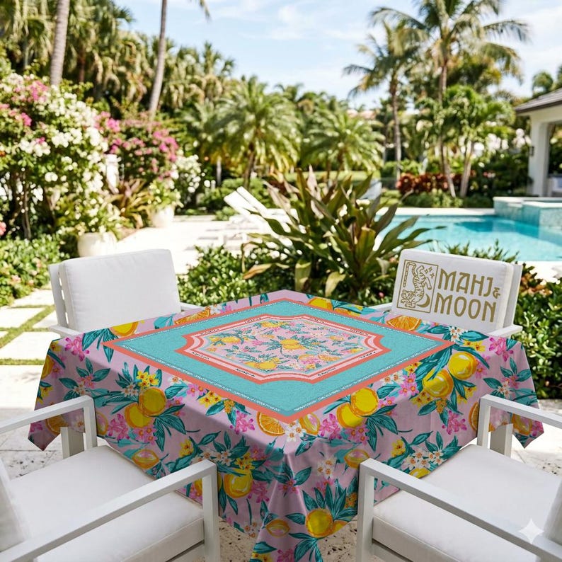 May include: A square tablecloth with a floral and citrus fruit pattern. The tablecloth has a light pink background with lemons, oranges, and flowers. A turquoise border frames a central design. The words "MAHJ & MOON" are on a pillow.