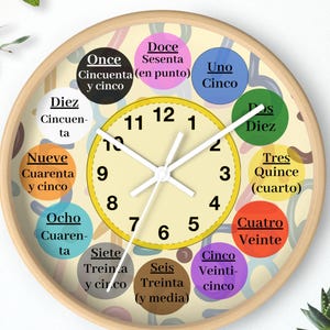 Puede incluir: Un reloj de pared redondo con marco de madera clara. La esfera del reloj es de color crema con números y manecillas negras. Círculos coloridos alrededor del borde muestran números en español. Una pequeña planta en maceta está debajo.