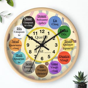 Puede incluir: Un reloj de pared redondo con marco de madera clara. La esfera es beige con círculos coloridos alrededor del borde, cada uno con un número en francés. Las manecillas son negras y el texto "Quelle heure est-il?" está en el centro.