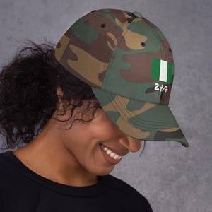 Puede incluir: Gorra de béisbol de camuflaje con la bandera nigeriana y el texto "24/7". La gorra tiene una visera curva y un diseño clásico de seis paneles. Los colores incluyen tonos de verde, marrón y negro.