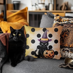 May include: A black cat with yellow eyes sits next to a decorative pillow. The pillow has a yellow background with black polka dots and a cartoon cat wearing a witch hat and sitting next to a carved pumpkin. The scene is set on a gray couch.