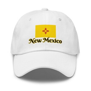 Puede incluir: Gorra de béisbol blanca con la bandera del estado de Nuevo México. La bandera es un rectángulo amarillo con un símbolo Zia rojo en el centro. Las palabras "New Mexico" están impresas en cursiva debajo.