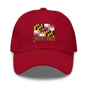 Puede incluir: Gorra de béisbol roja con el diseño de la bandera del estado de Maryland. El diseño de la bandera incluye un patrón negro y dorado, y un patrón de cruz blanco y rojo. El texto "your text" está debajo.