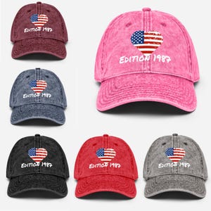 Puede incluir: Seis gorras de béisbol en varios colores: burdeos, rosa, azul, negro, rojo y gris. Cada gorra presenta un diseño de bandera americana en forma de corazón y el texto "EDITION 1987" en blanco.