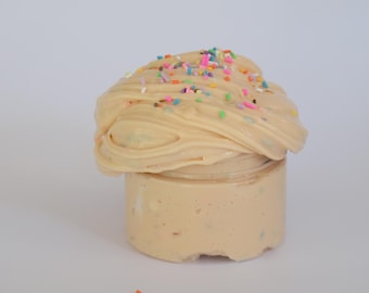 Puede incluir: Un tarro de slime beige cubierto con chispas de colores. El slime tiene una textura suave y cremosa y está contenido en un tarro de plástico transparente. Las chispas son de varios colores, incluyendo rosa, verde y amarillo.