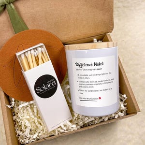 Può includere: Una confezione regalo contenente una candela bianca con coperchio in legno, una scatola di fiammiferi e un sottobicchiere rotondo. La candela ha il testo "Difference Maker" e una definizione. La scatola è legata con un nastro verde.