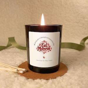 Op de afbeelding: Een brandende zwarte kaars met een wit etiket met de tekst "Eid Mubarak" in het rood. De kaars staat op een bruine onderzetter, met lucifers ernaast. Een groen lint is op de achtergrond.