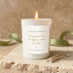 May include: A lit white candle with a gold flame, featuring a label with the text "DEAR MRS. ROBINSON, Thank you for being the difference maker in my life, With love from Oliver Smith." Two matches are next to the candle.