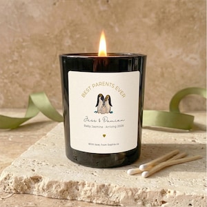 May include: A lit black candle with a white label that reads "Best Parents Ever" with a penguin illustration. The text "Jess & Damian" and "Baby Jasmine - Arriving 2026" are also on the label. Matches and a green ribbon are nearby.
