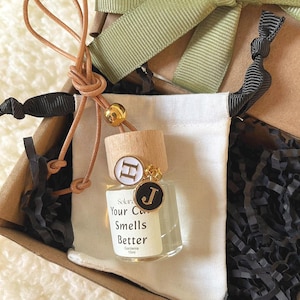 Könnte beinhalten: Kleine Glasflasche mit Holzstopfen und Lederschnur in einer Geschenkbox. Auf der Flasche steht "Your Car Smells Better". Der Stopfen ist mit einem goldenen Anhänger und dem Buchstaben "H" verziert. Die Box ist mit einem grünen Band dekoriert.