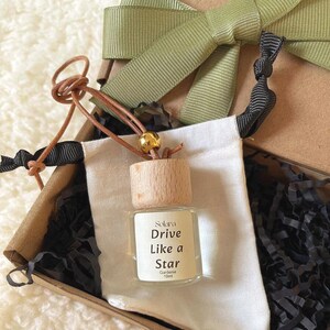 Può includere: Una piccola bottiglia di vetro con tappo in legno e cordino in pelle, presentata in una confezione regalo. L'etichetta della bottiglia recita "Drive Like a Star" e "Gardenia 10ml". La scatola è legata con un nastro verde.