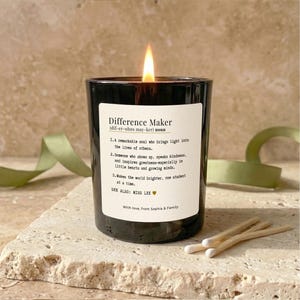 Könnte beinhalten: Eine brennende schwarze Kerze mit einem weißen Etikett mit der Aufschrift "Difference Maker". Das Etikett enthält eine Definition und eine Nachricht. Zwei Streichhölzer liegen neben der Kerze. Ein grünes Band ist im Hintergrund.