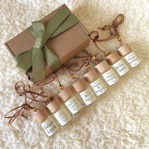 May include: A gift set featuring small glass bottles with wooden caps and leather cords. Each bottle has a label with a different positive message, such as "Good Luck" and "Drive Safe." The set includes a brown gift box tied with a green ribbon.