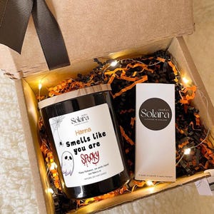 Puede incluir: Una caja de regalo con temática de Halloween que contiene una vela negra con tapa de madera y una etiqueta blanca que dice "Hanna Smells like you are Spooky". La caja también incluye una caja blanca con el logotipo de Solara Candles y papel naranja y negro.