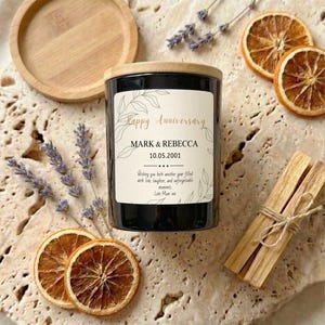 May include: A black candle with a wooden lid, labeled "Happy Anniversary" with the names Mark & Rebecca and the date 10.05.2001. The candle is surrounded by dried orange slices, lavender sprigs, a wooden coaster, and a bundle of palo santo sticks.