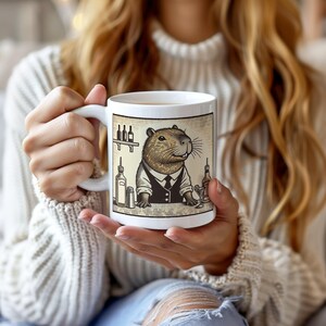May include: White ceramic mug with a graphic of a capybara wearing a bartender's outfit and holding a cocktail shaker. The capybara is standing behind a bar with bottles of liquor and a shaker.