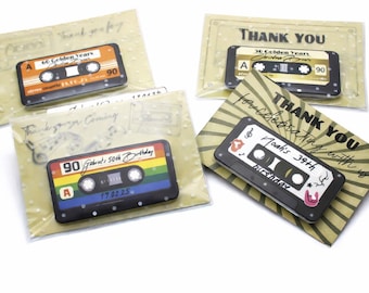 May include: Four rectangular party favors featuring cassette tape designs on printed cards. One orange tape says 60 Golden Years, one gold tape says 30 Golden Years, one rainbow tape says Gabriel's 50th Birthday, and one black tape says Noah's 39th birthday.