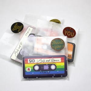 May include: Clear cellophane bags with various retro-themed designs. Each bag contains a cassette tape design, with the text "90 Emily and Ramiro" and "Thank You" stickers. The bags are sealed with round stickers.