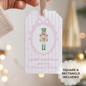 May include: A Christmas gift tag with a pink and white striped background. The tag features a nutcracker illustration in an oval frame with a bow at the top. The text reads "MERRY CHRISTMAS from the Anderson Family". A cream ribbon is attached.